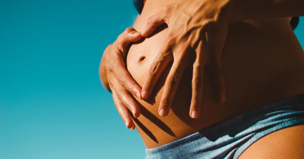 Kobieta w ciąży układa dłonie w kształt serca na brzuchu na tle niebieskiego nieba