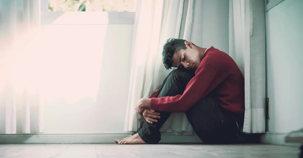 Smutny młody mężczyzna siedzi pod oknem i trzyma głowę na kolanach