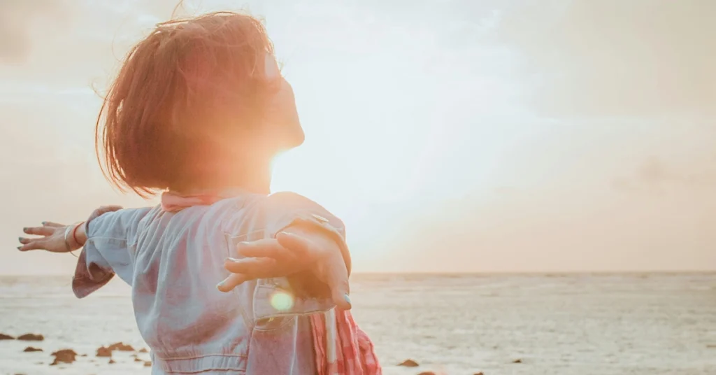 Kobieta z rozłożonymi szeroko rękami stoi na plaży na tle zachodzącego słońca
